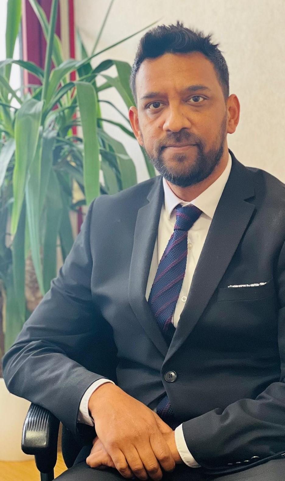Professional headshot of a bearded man in a dark suit and navy tie, seated with a plant in the background